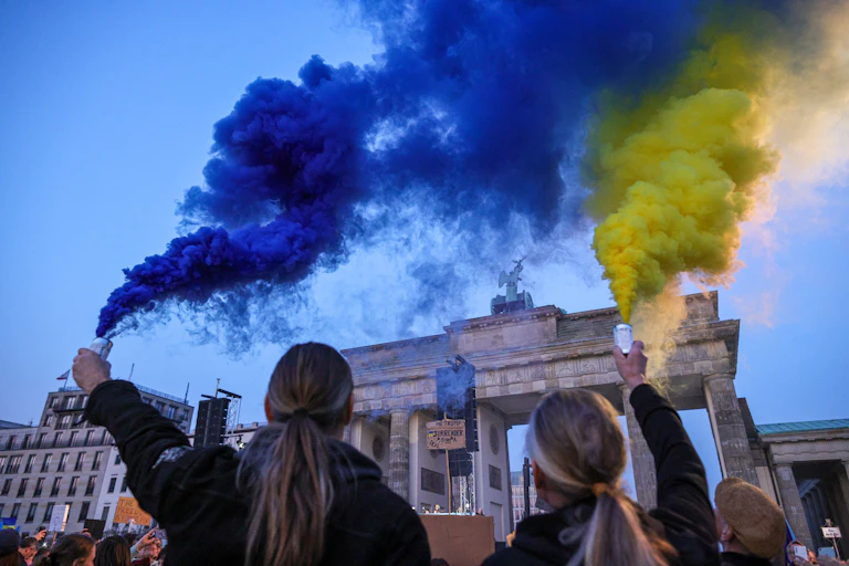 Two people hold flares: one emiting blue smoke, the other yellow.