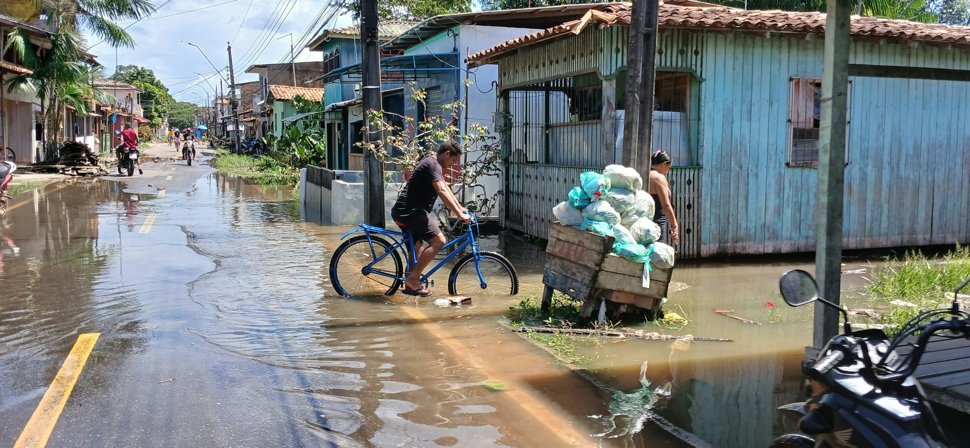 Foto mostra casas sobre palafitas e rua tomada de água por onde passa uma bicicleta