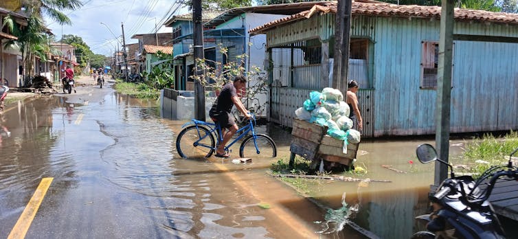 Foto mostra casas sobre palafitas e rua tomada de água por onde passa uma bicicleta