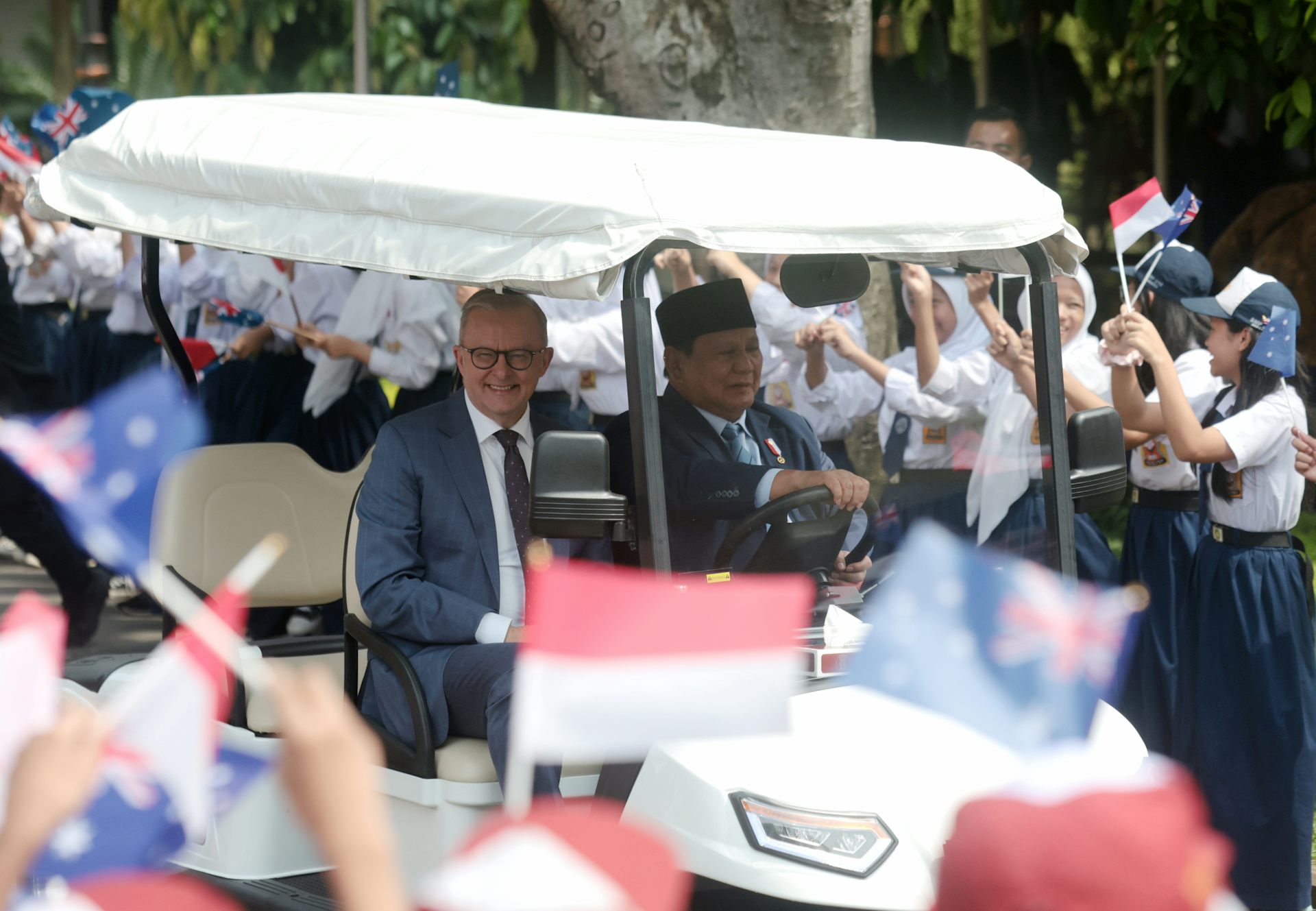 Anthony Albanese and Prabowo Subianto drive in a golf cart among smiling people waving flags.