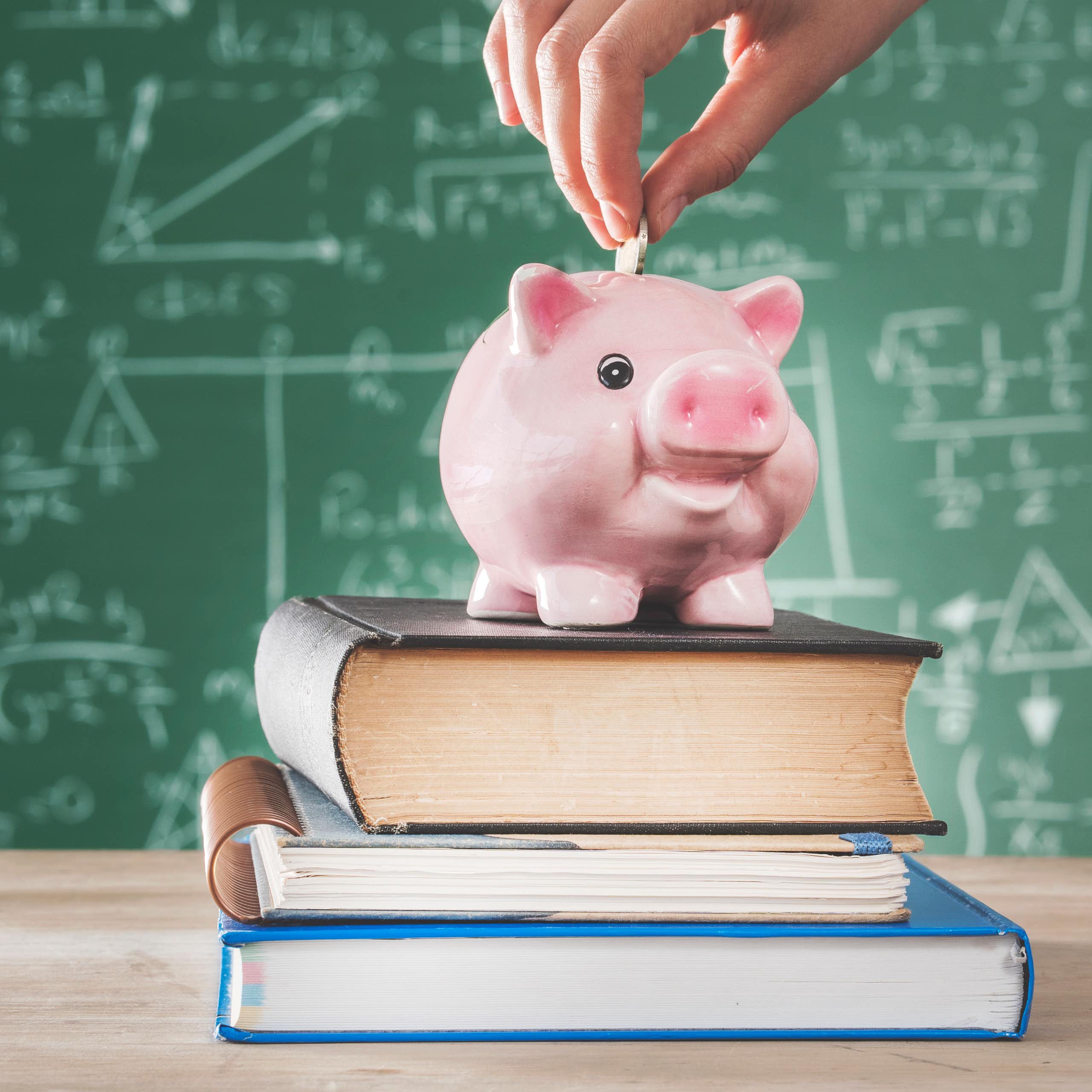 Female putting coin into piggy bank on a stack of text books in front of a blackboard
