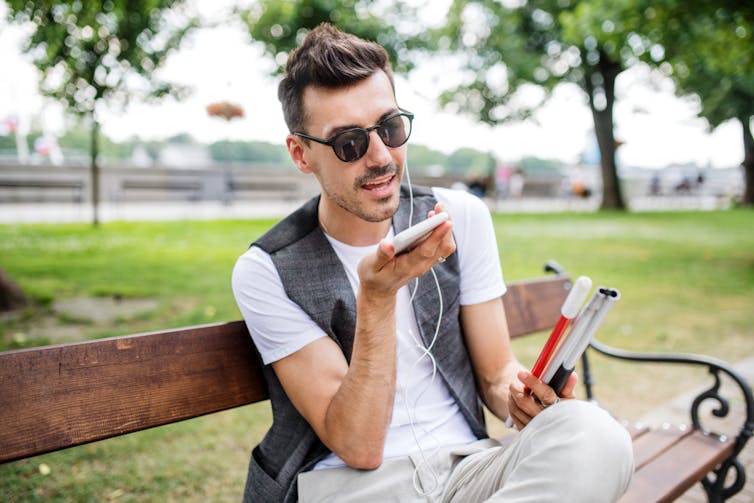A young man sits on a park bench with phone and headphones in hand, holding a folded white cane.