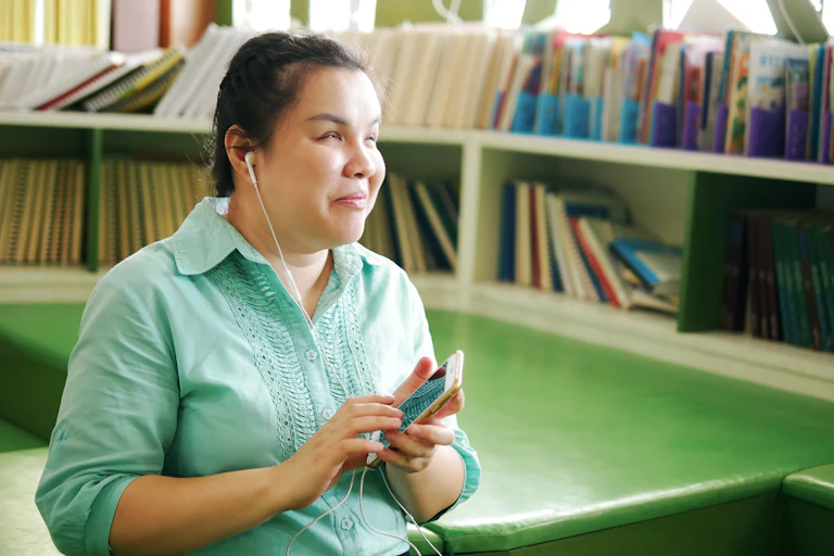 A young blind woman sits in a sunny library listening to a smartphone with headphones plugged in.