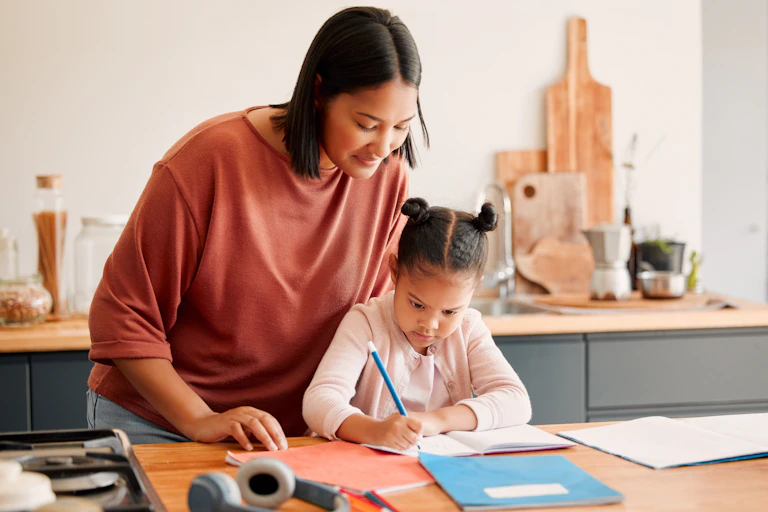 Mother stands behind her child, who is concentrating on writing, in the kitchen