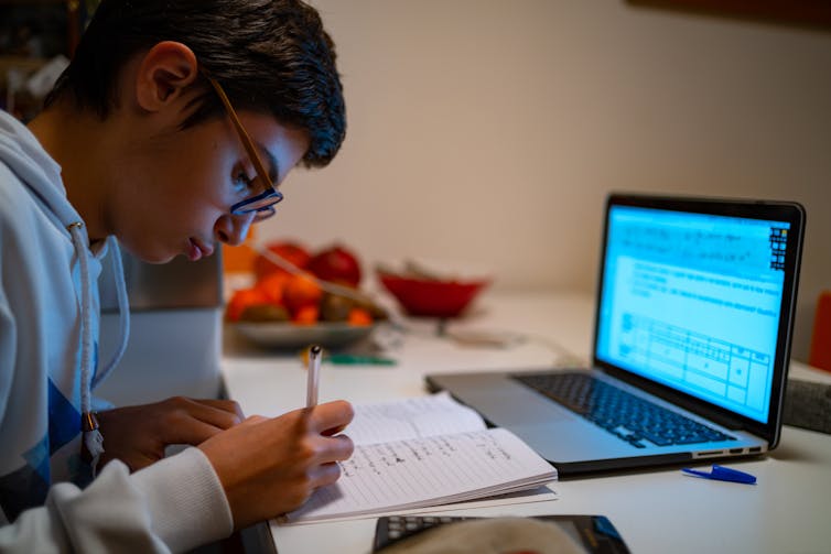 A young man writes in front of a laptop.