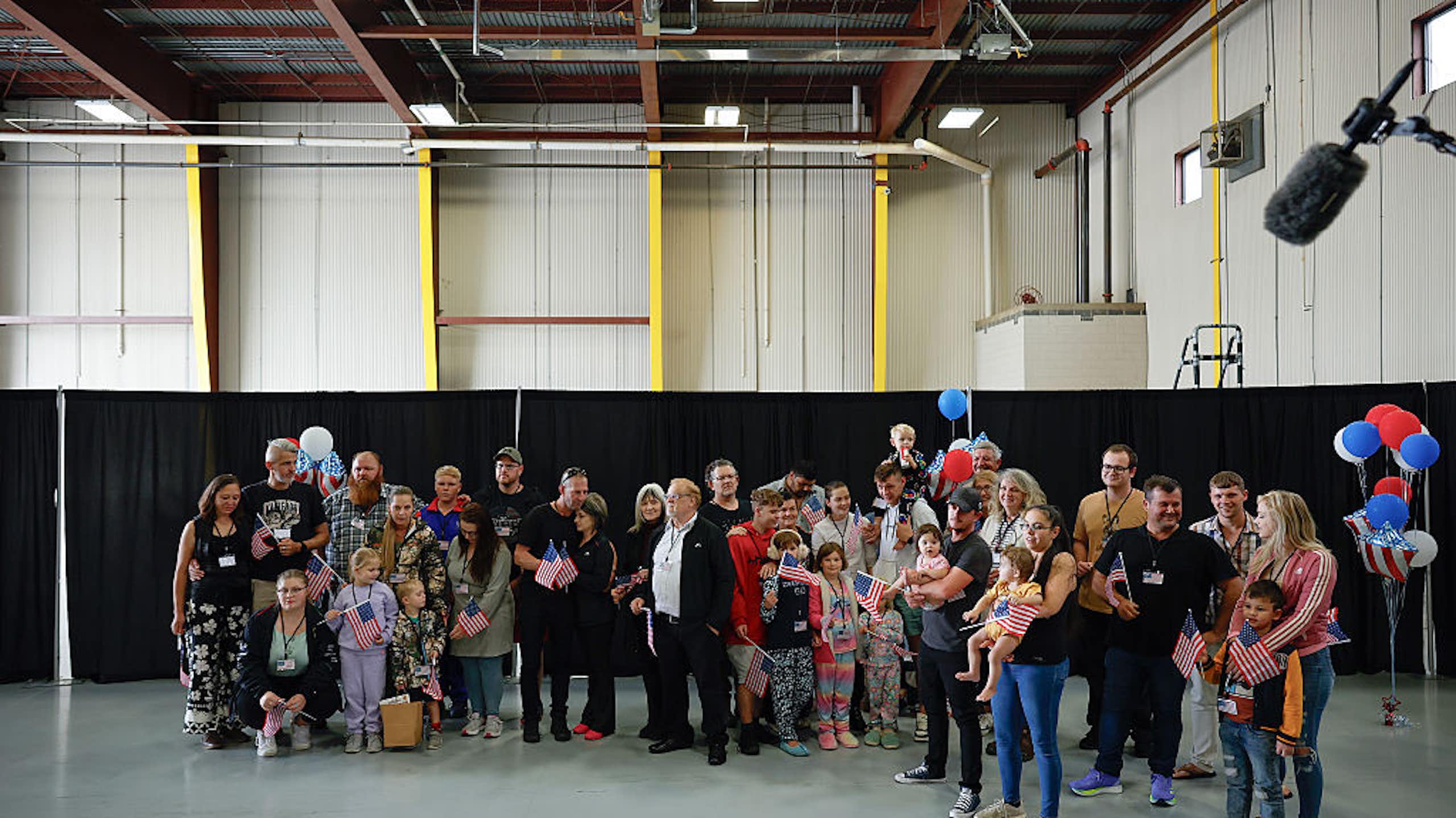 A group of adults and children standing in a large indoor space, with some balloons in the background. Some are holding small US flags.