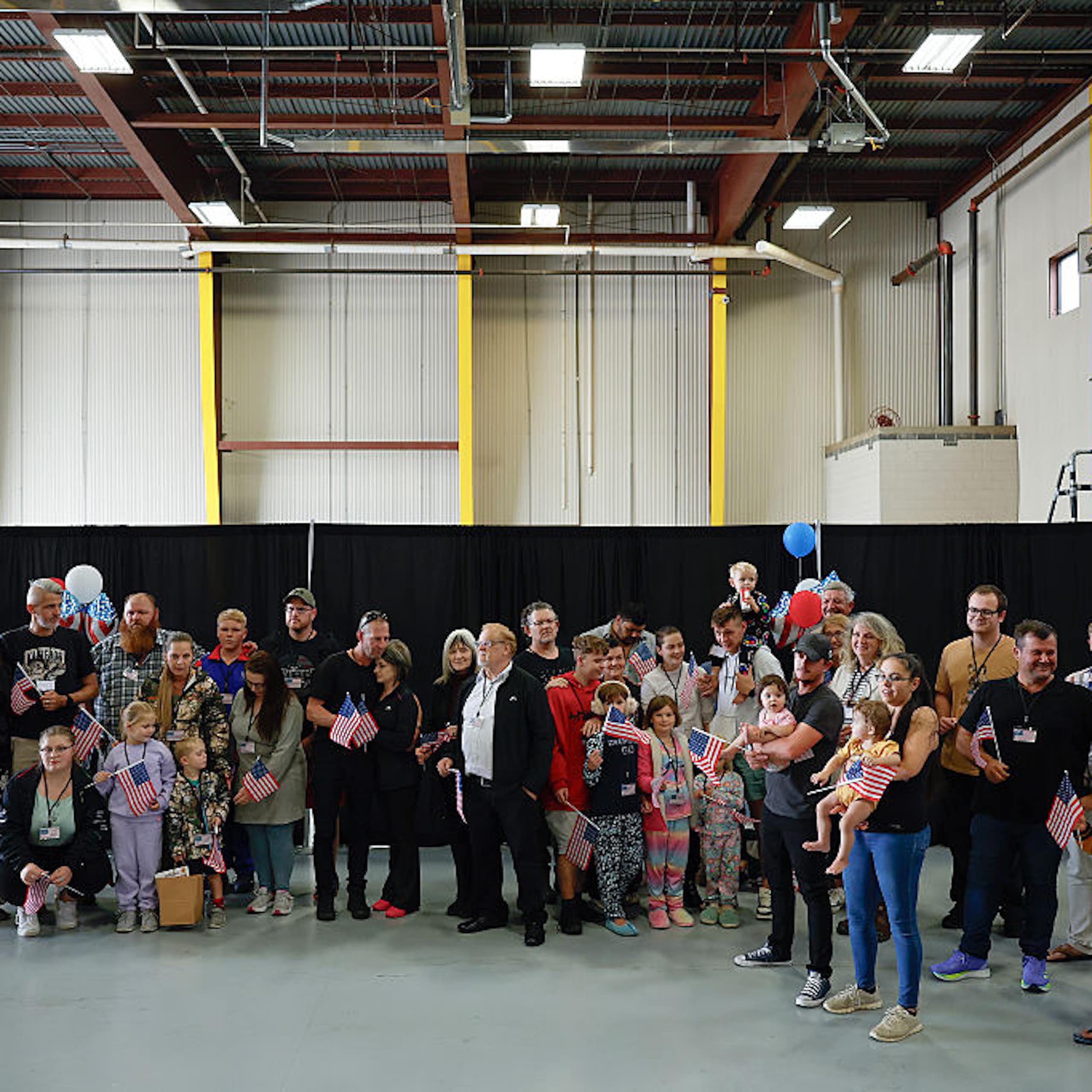 A group of adults and children standing in a large indoor space, with some balloons in the background. Some are holding small US flags.