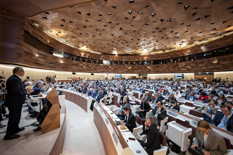 In a cavernous hall, a large assembly of people watch a speaker on stage delivering a report.