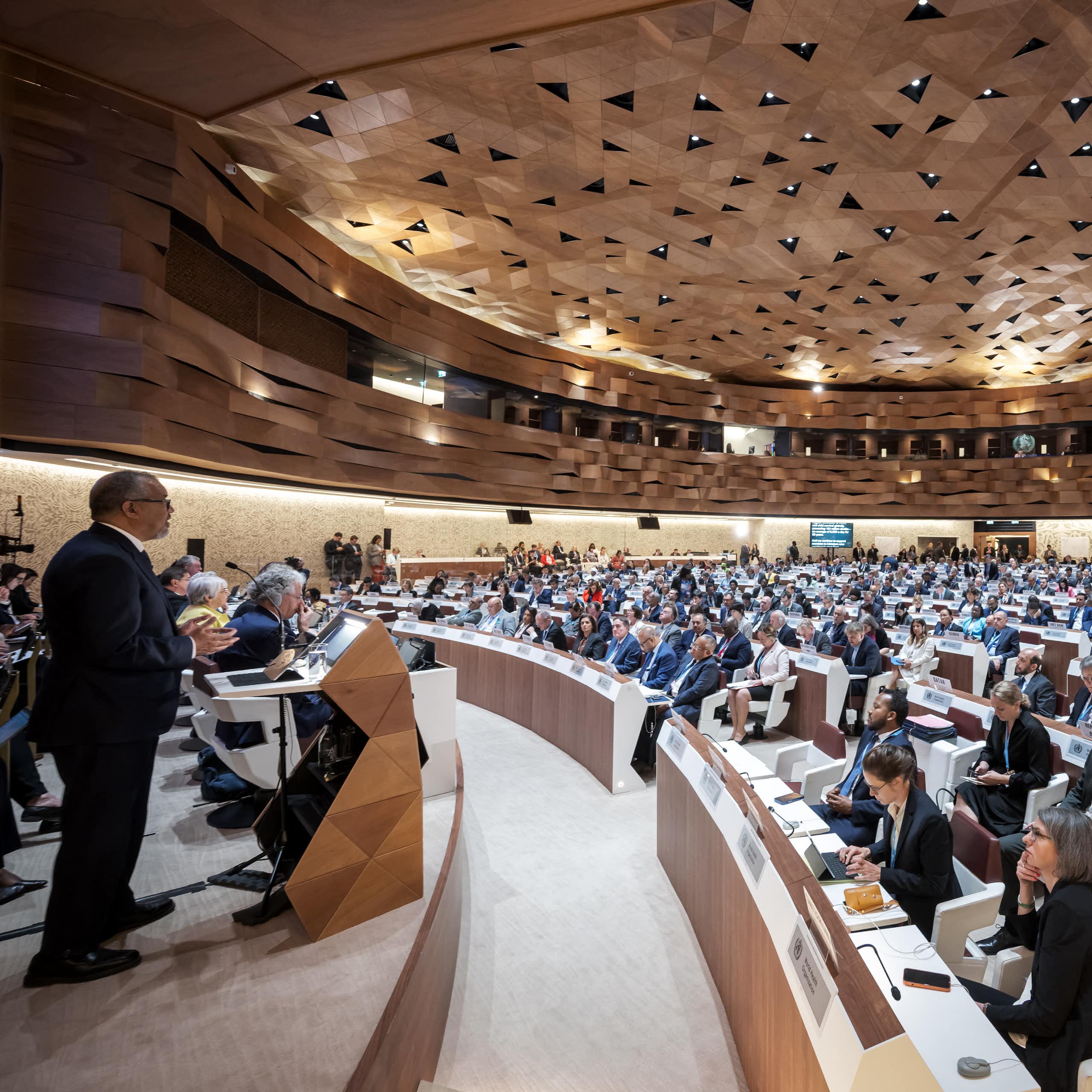 In a cavernous hall, a large assembly of people watch a speaker on stage delivering a report.