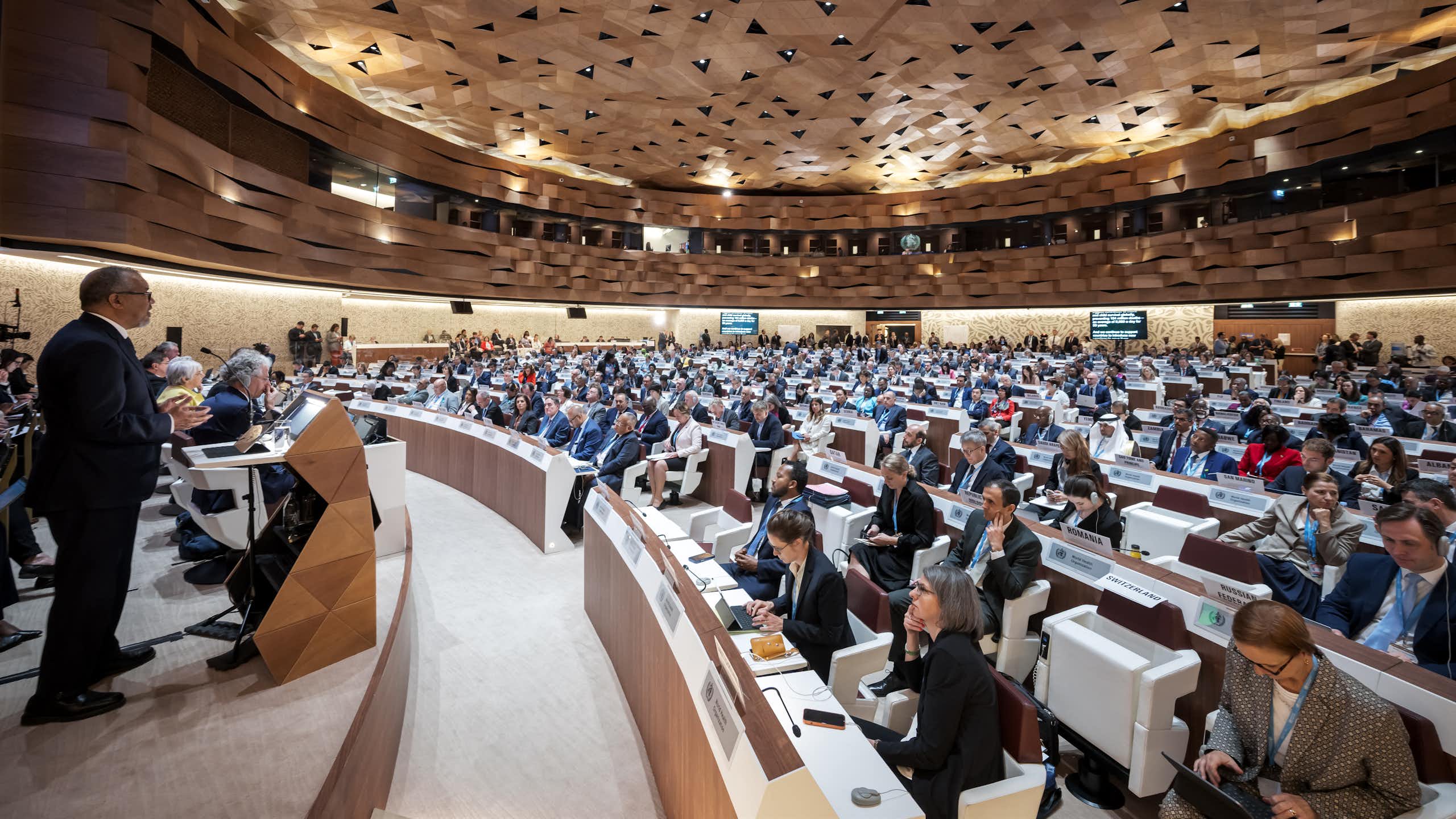 In a cavernous hall, a large assembly of people watch a speaker on stage delivering a report.
