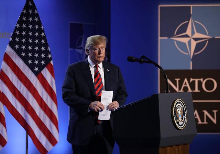 A man with light yellow hair and a black suit and red tie stands at a podium with an American flag behind him and a sign that says NATO next to the podium.