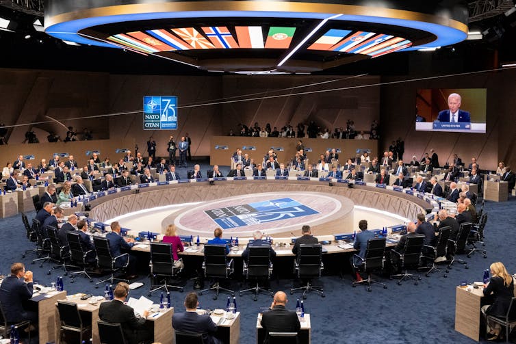 People sit around a large, circular table in a large room that has flags of different countries projected above them.