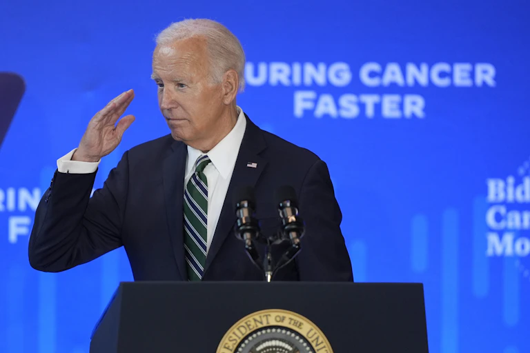 Joe Biden with hand partially raised in what seems like a salute, standing behind a lectern with a backdrop reading 'CURING CANCER FASTER'