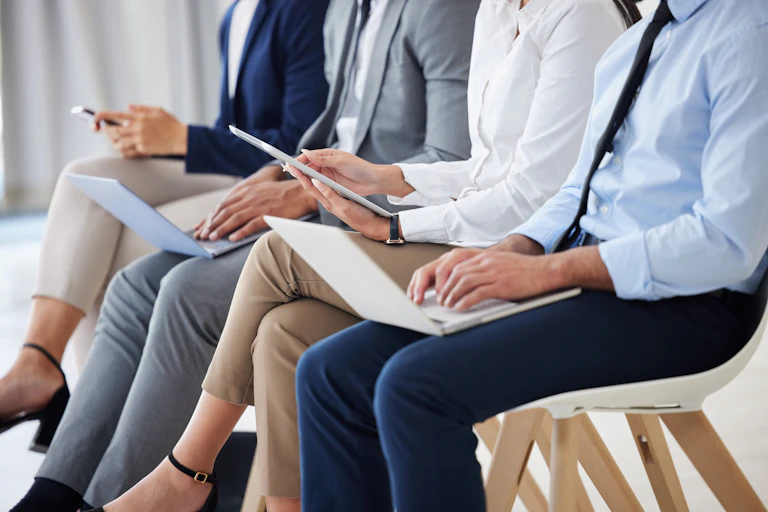 Four people in business attire, seen from the neck down, sit side-by-side on chairs