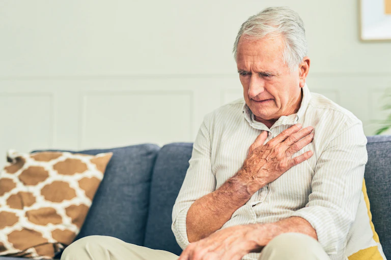 A man sitting on a couch appears to be in pain as he holds his hand over his chest.