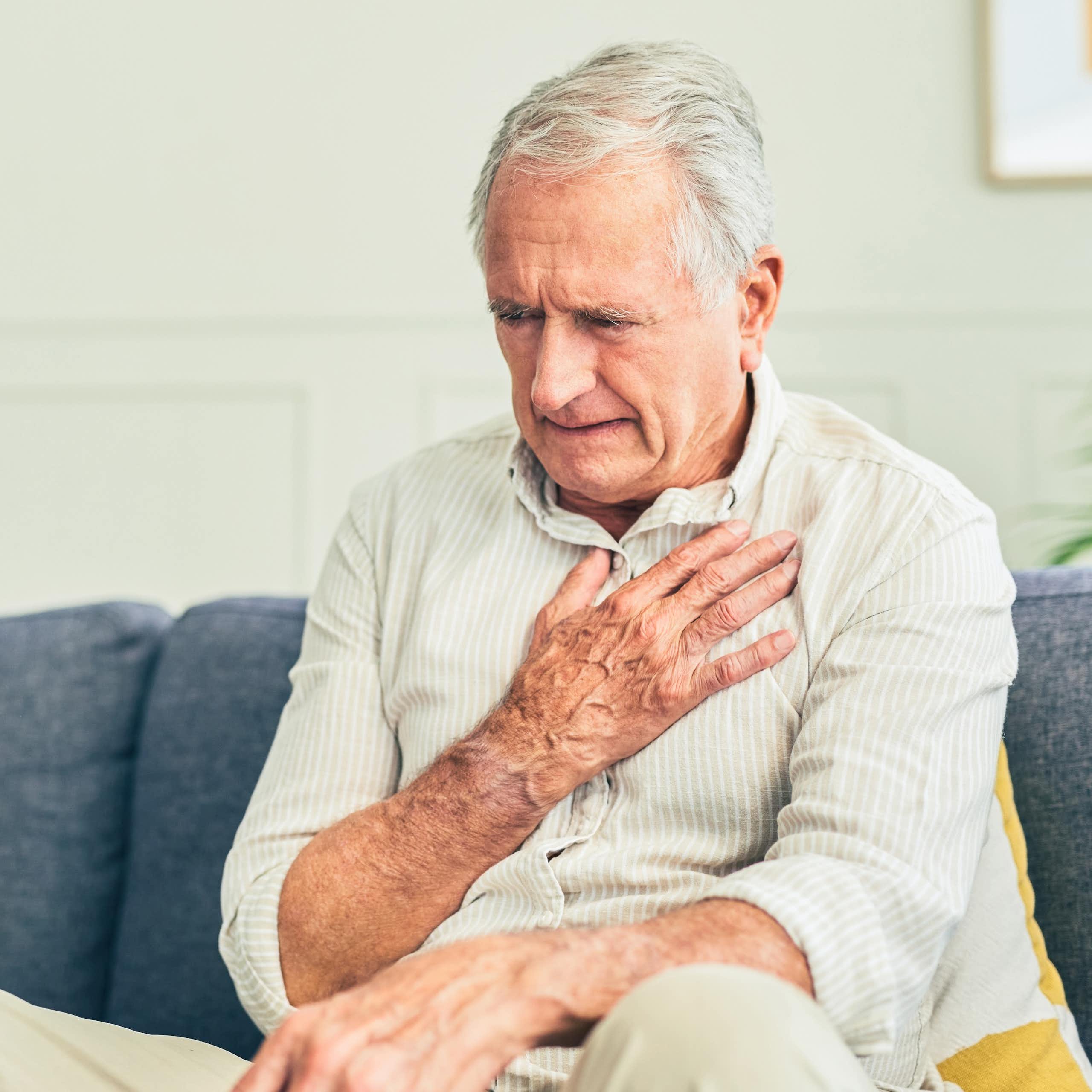 A man sitting on a couch appears to be in pain as he holds his hand over his chest.