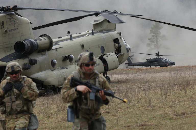 Two men in camouflage stand in front of two helicopters on the ground on a cloudy day.