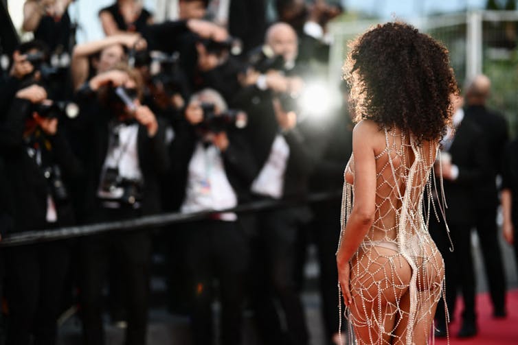 Young woman with curly hair and a skimpy beaded dress poses on the red carpet in front of a crowd of photographers.