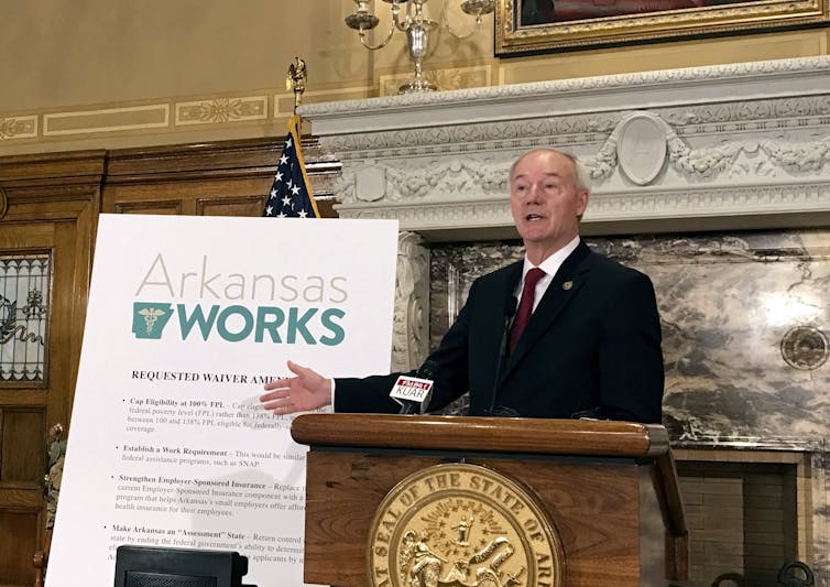 Man at lectern in a suit, next to a sign saying Arkansas Works