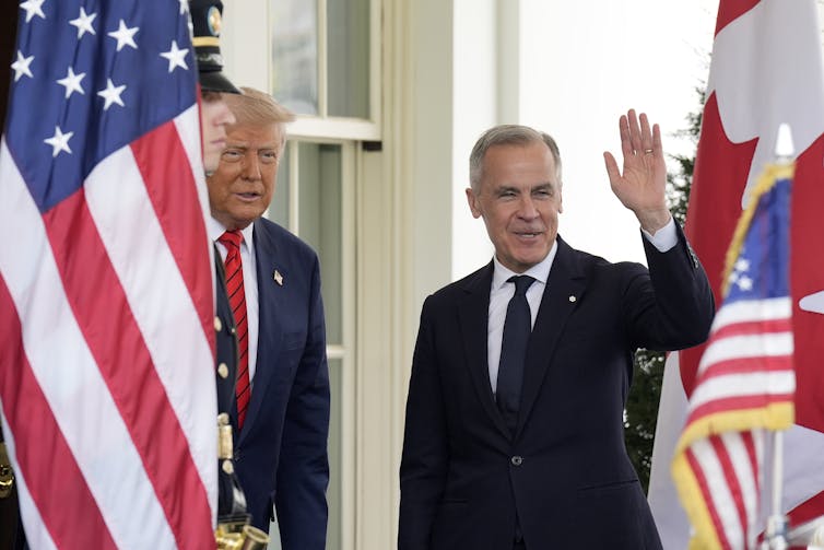 Un hombre con una sonrisa de cabello gris corto y saluda al lado de la bandera canadiense, mientras que un viejo hombre de la rotonda con cabello gris azulado y bronce naranja se encuentra detrás de la bandera estadounidense.
