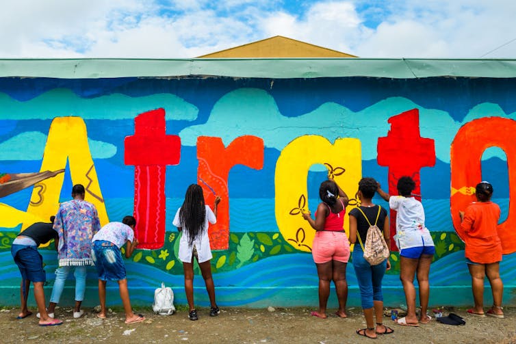 About half a dozen girls, seen from behind, paint a brightly colored mural on the wall of a blue building.