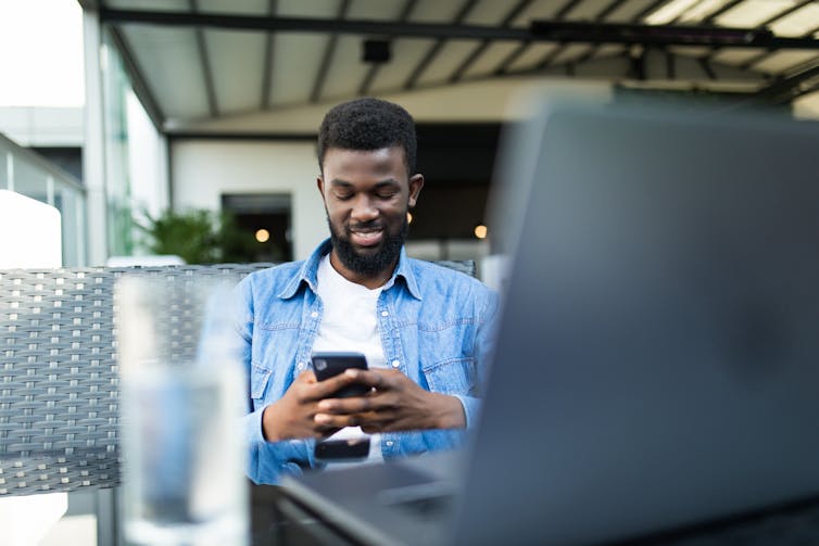 The man smiles at the phone with an open laptop.