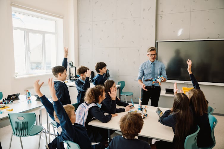 A small group of children put their hand up in class. A teacher stands close to them in front of a board.