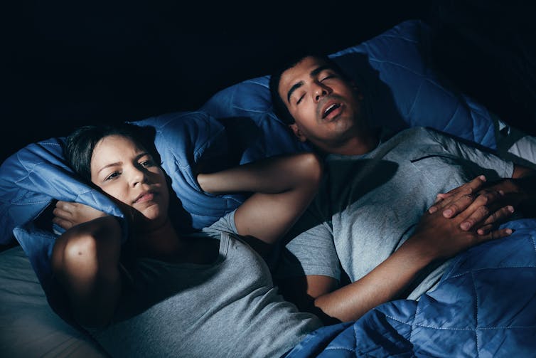 A couple in bed. The woman holds her pillow to her ears while her partner sleeps with his mouth open.