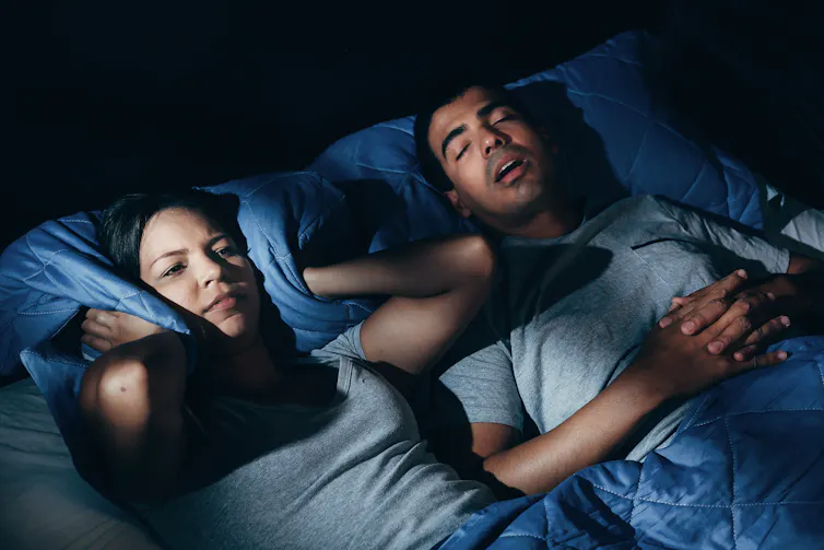 A couple in bed. The woman holds her pillow to her ears while her partner sleeps with his mouth open.