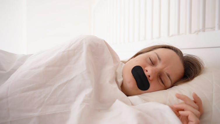 A woman asleep in a bed with black tape over her mouth.