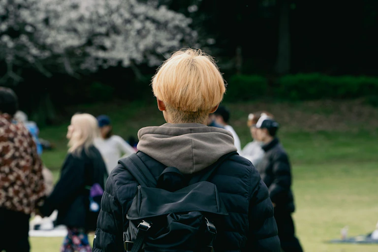 A young person with a back pack faces a small crowd of people in a park area.