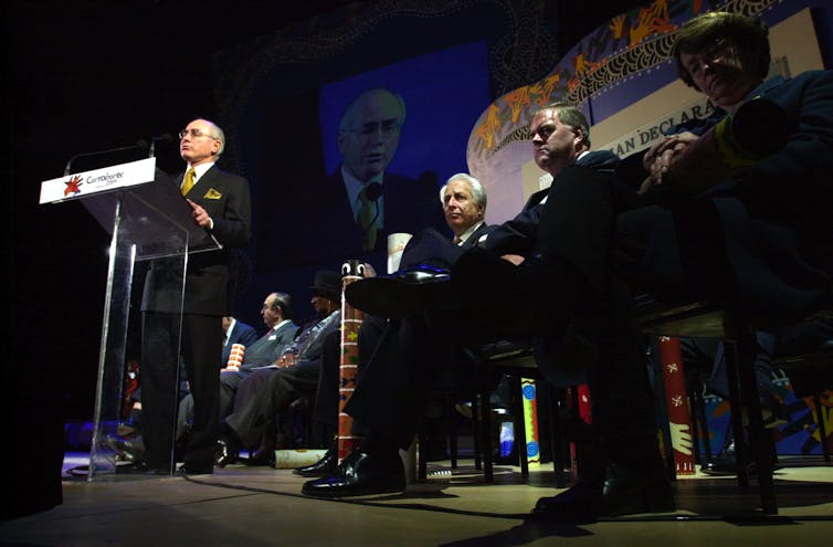 John Howards stands at a lectern on a stage in front of other men sitting down