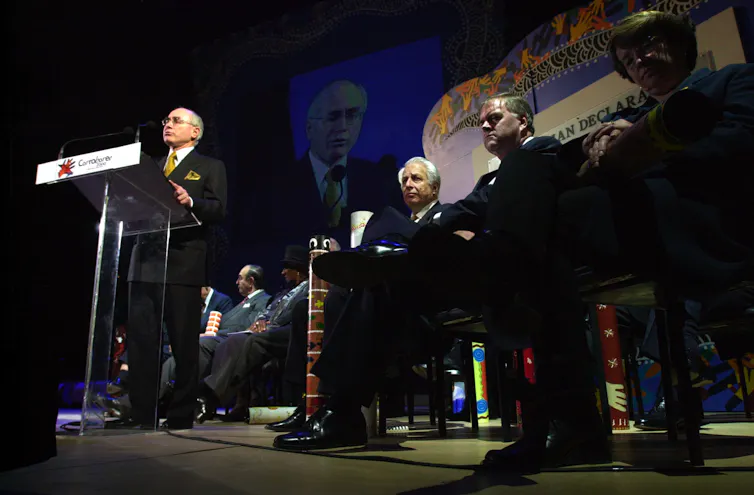 John Howards stands at a lectern on a stage in front of other men sitting down