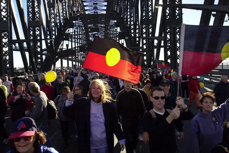 A large group of people march over the Sydney Harbour Bridge waving Aboriginal flags