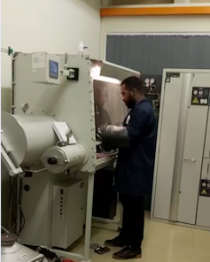 A man stands in front of a giant machine taller than his head with his hands inside a silver tube