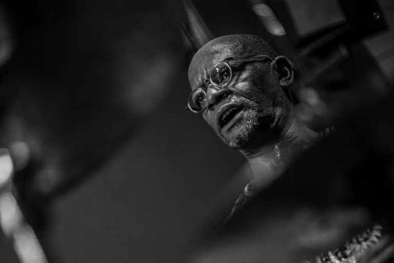 A black and white photo of an African man with a bald head wearing glasses, concentrating, his mouth open.