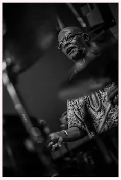 A black and white photo of an African man in glasses and a traditional shirt sweating under the lights as he drums, mouth open.