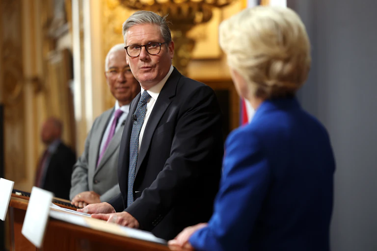 view from the side of Costa, Starmer and von der Leyen standing at podiums. Starmer's face is in focus and he is turned towards von der Leyen.