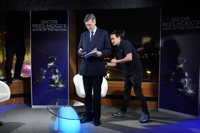 Rees-Mogg checks his phone while standing in the middle of a soundstage, in front of a banner for his show Jacob Rees-Mogg's State of The Nation
