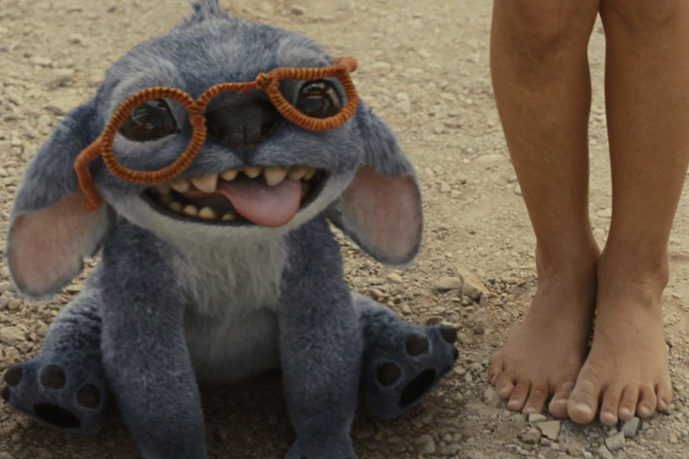 A small alien-like creature that looks a bit like a dog grinning at the camera next to a pair of child's legs.