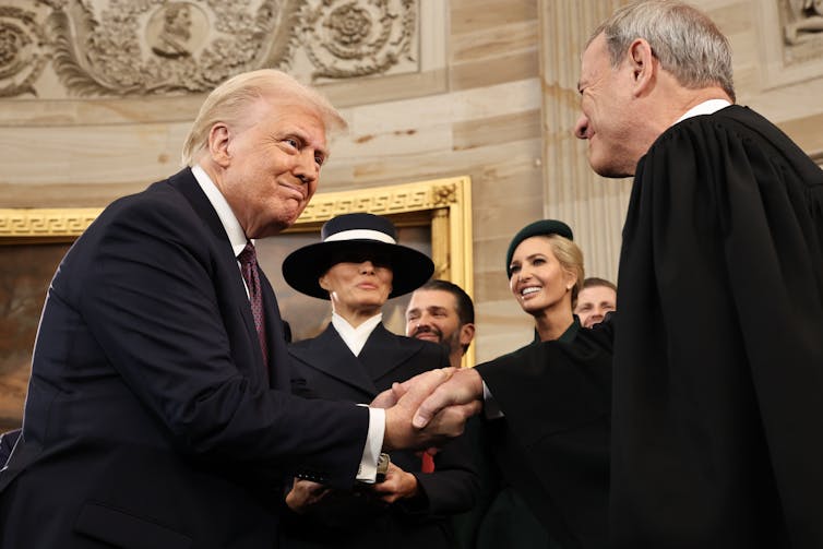 Two white men, one wearing a black blazer and the other wearing a black robe, shake hands and look at each other and smile. Two women and one man dressed formally stand smiling near them.