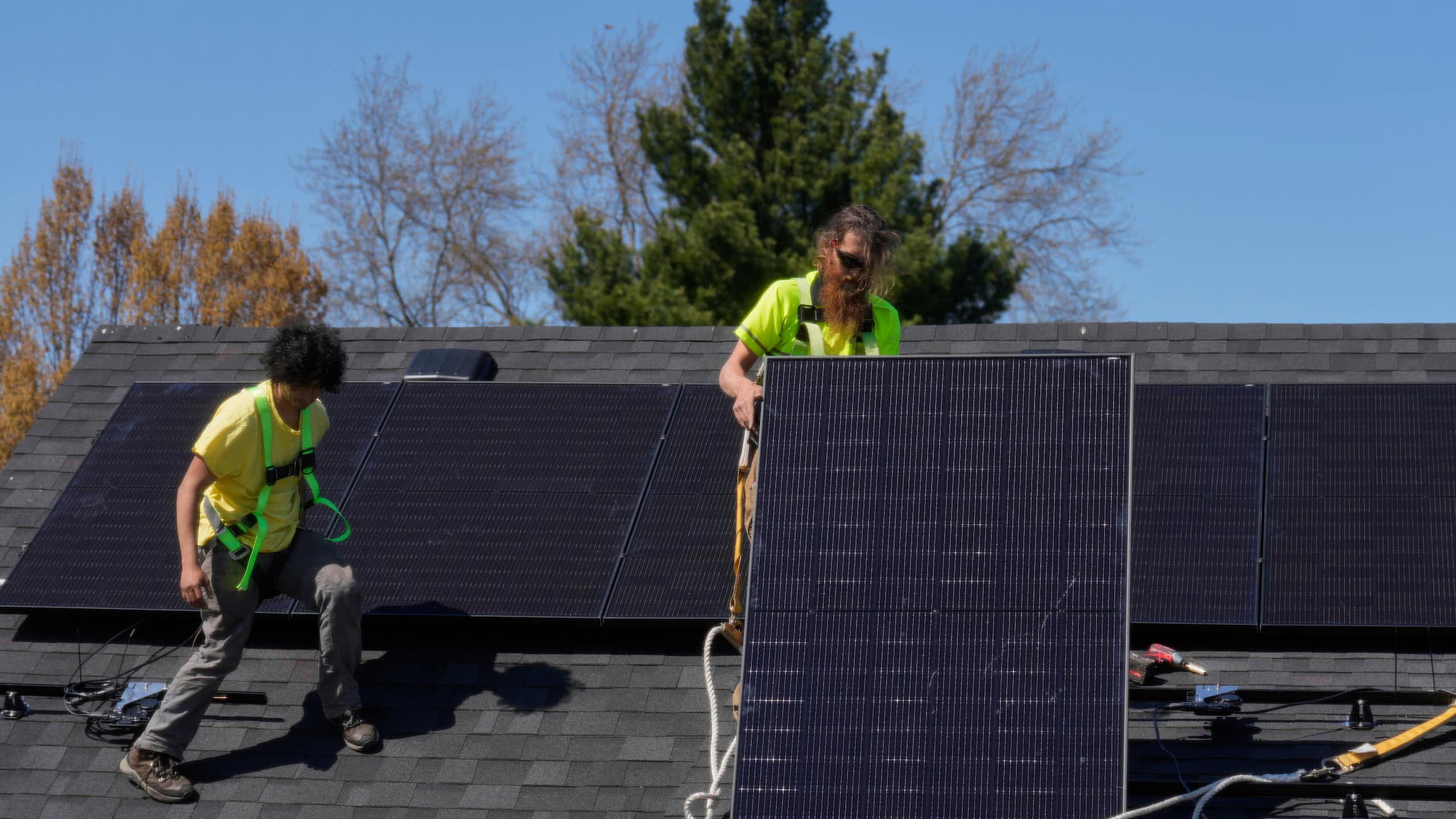 Two people move solar panels around on a rooftop.