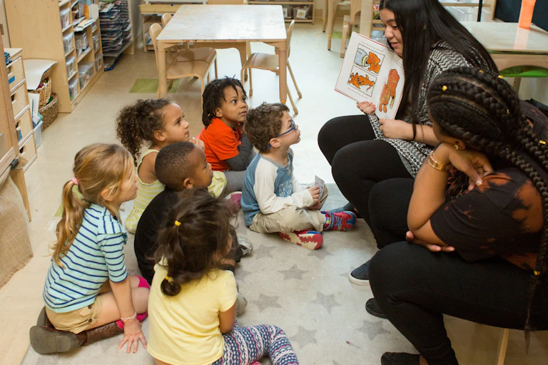 Two adults on chairs, one with a book, in front of a circle of young children.