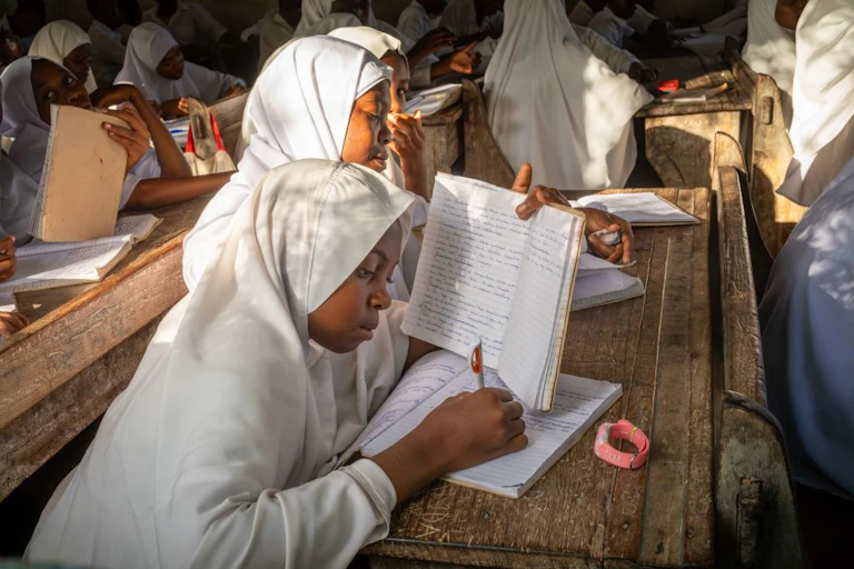 Girls in white robes and headscarves sit at a desk in a school classroom, writing in exercise books