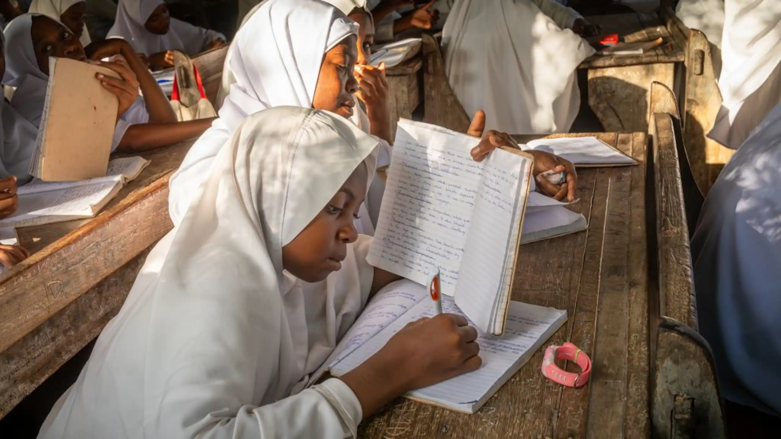 Girls in white robes and headscarves sit at a desk in a school classroom, writing in exercise books