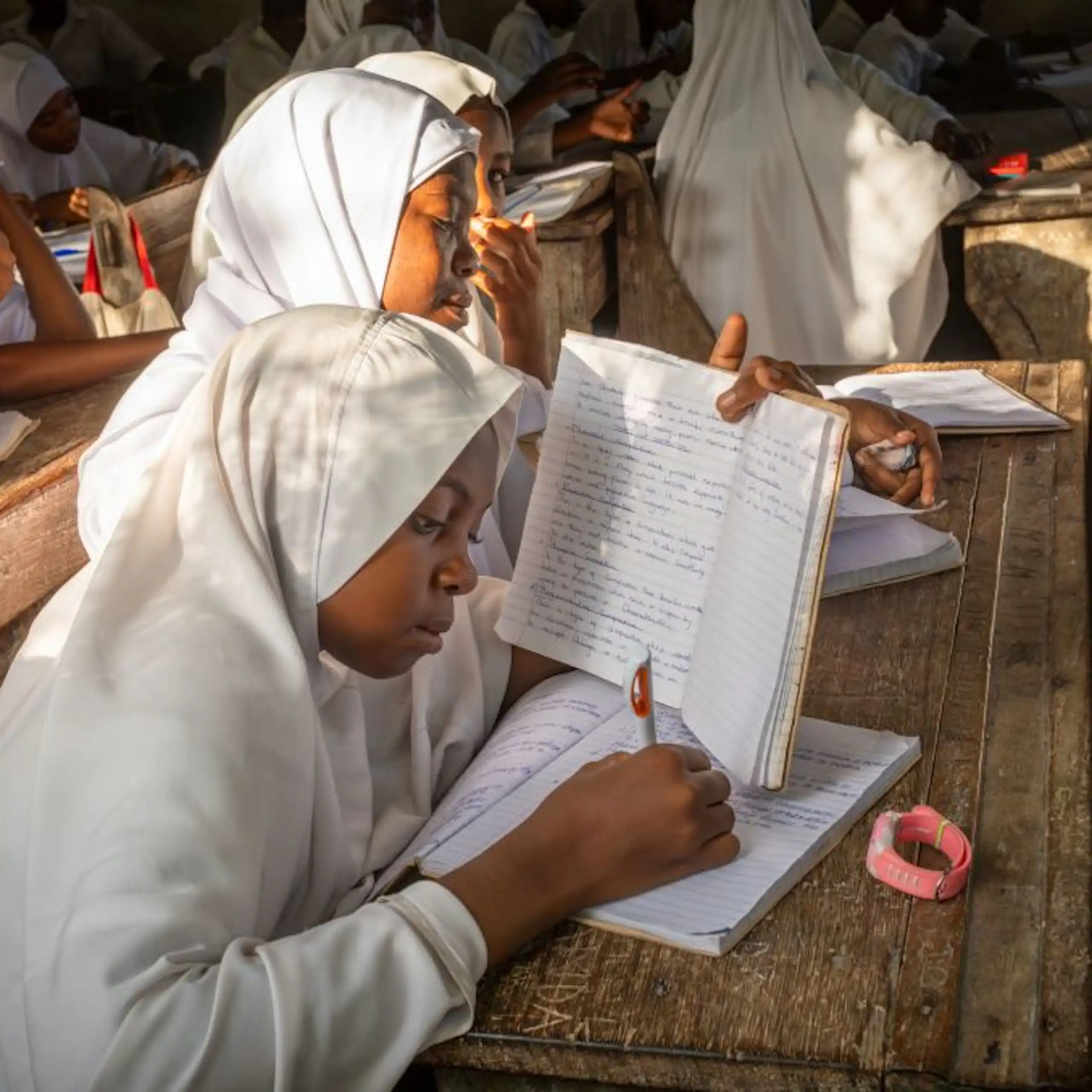 Girls in white robes and headscarves sit at a desk in a school classroom, writing in exercise books