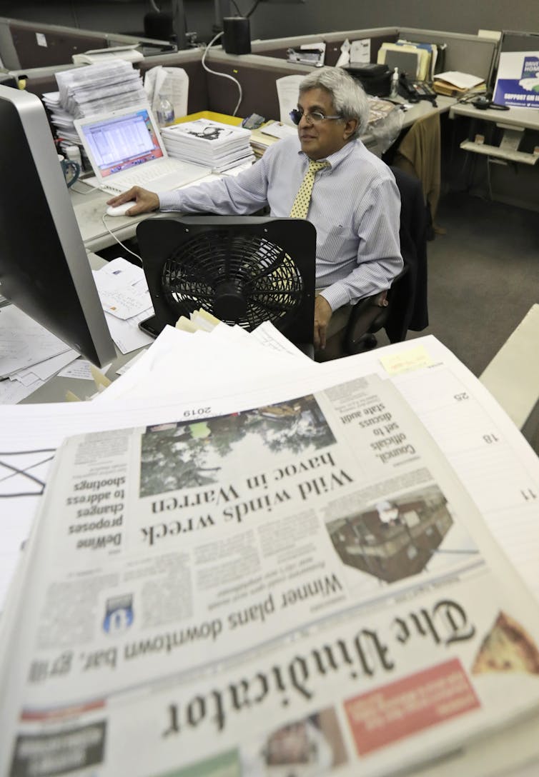 A newspaper on a table with a man in a shirt and tie behind it.