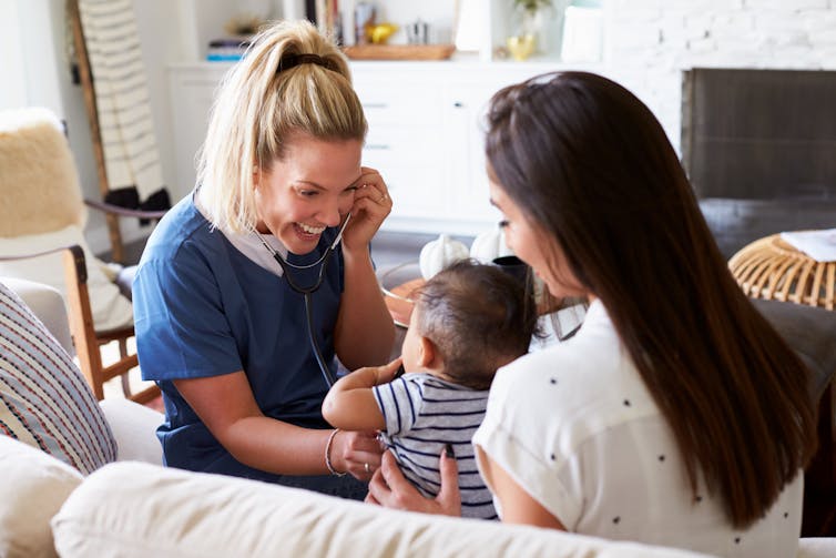 A woman in blue scrubs holding a stethoscope to an infant held by a young woman seen from behind