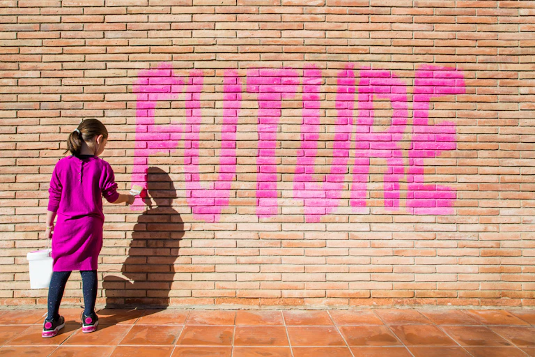 A young girl, dressed in a pink dress, paints the word, 'Future,' on a brick wall.