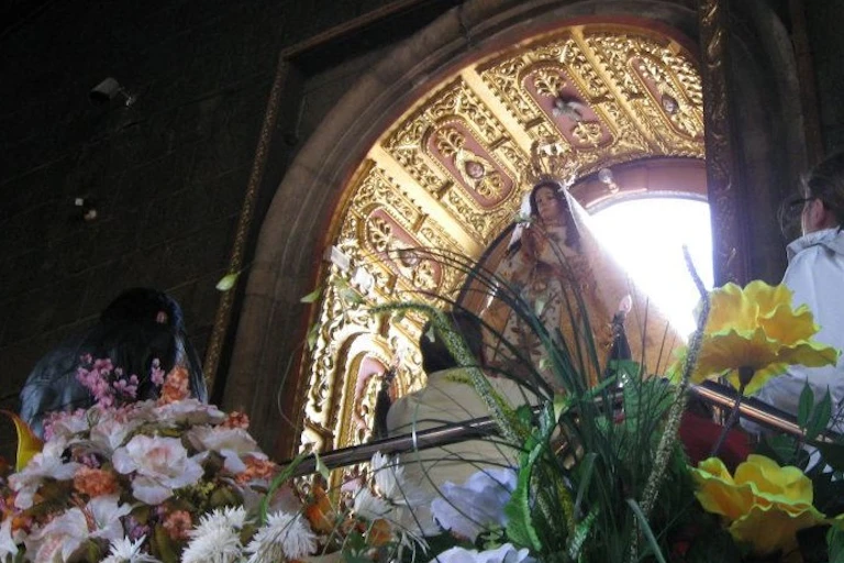 An elaborately decorated image of the Virgin Mary stands within an ornate enclosure, adorned with flowers placed at its base.